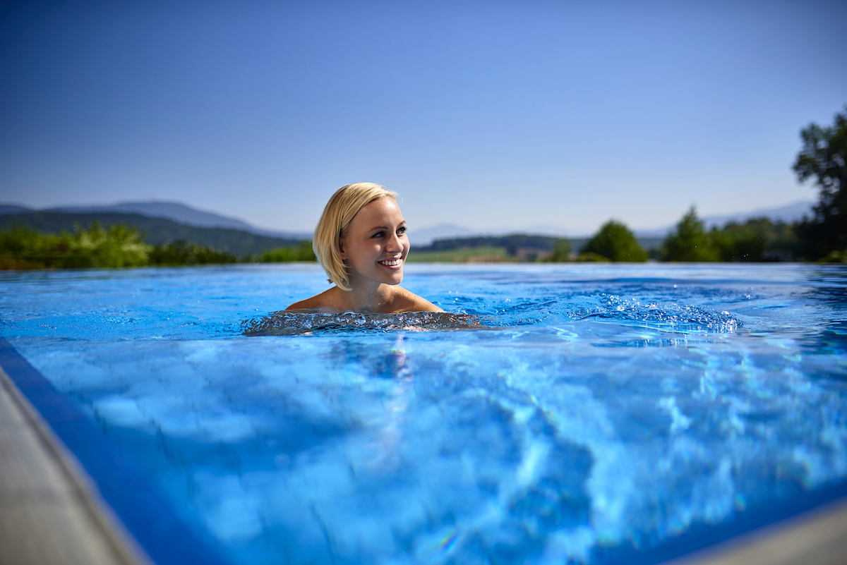 Außenpool mit lächelnder Frau im Wasser, Panoramablick auf bewaldete Hügel und blauen Himmel im Hintergrund