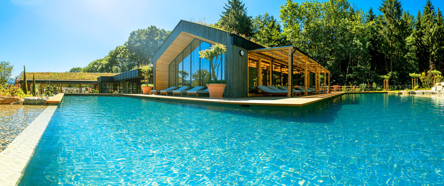 Großer Natur-Pool mit moderner Wellness-Architektur, umgeben von grünen Bäumen und blauem Himmel, mit Holz- und Glaspavilions am Ufer