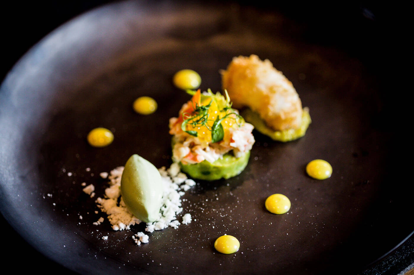 Fein angerichtete Vorspeise mit Cremetupfen und Deko auf dunklem Teller